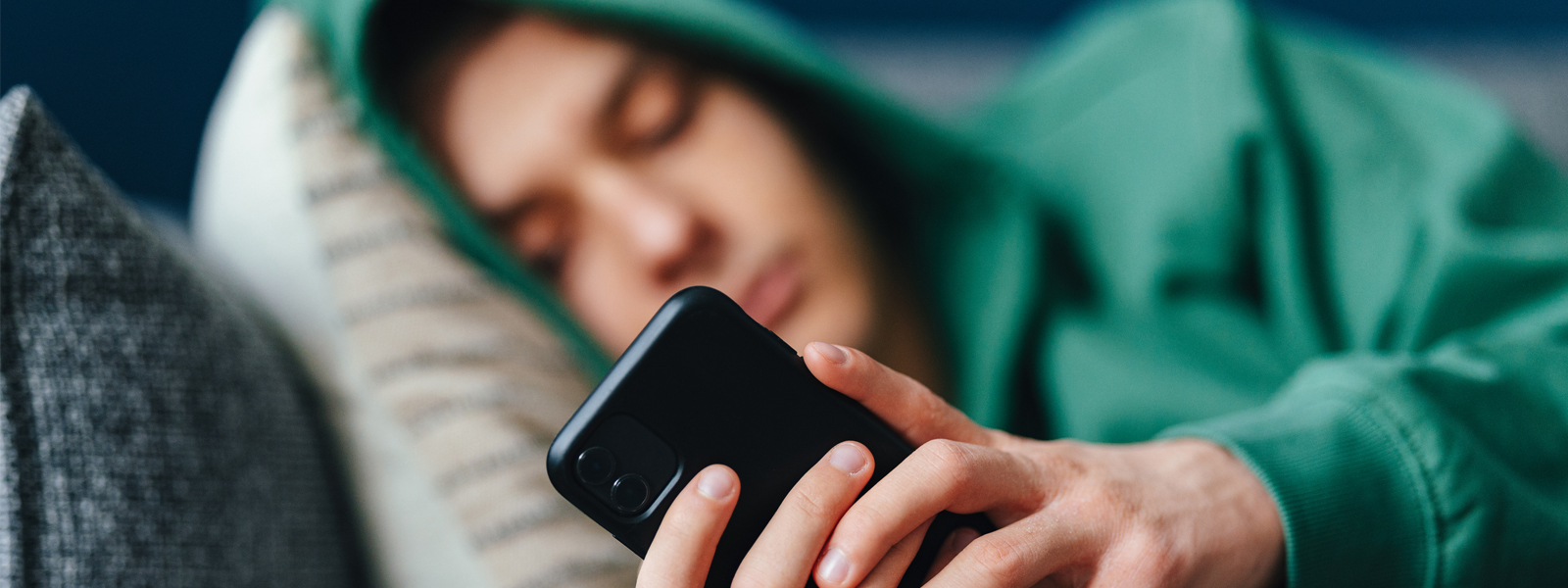 teen laying on couch with green hoodie on head, holding and looking sadly at phone.